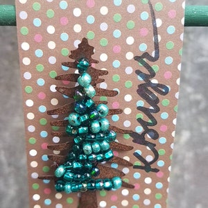 Puede incluir: Un adorno de &aacute;rbol de Navidad de cuentas azul turquesa sobre una tarjeta marr&oacute;n con lunares blancos. La tarjeta tiene la palabra "Forever" escrita en negro.