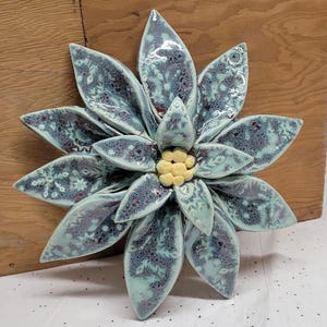 May include: A ceramic flower sculpture with a glossy finish. The petals are a teal color with a speckled pattern and snowflake designs. The center of the flower is yellow. The sculpture is displayed on a wooden surface.