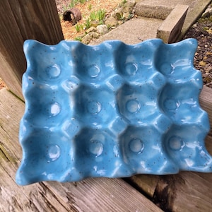Puede incluir: Un soporte para huevos de cerámica turquesa vibrante con detalles moteados. El soporte tiene un diseño ondulado y capacidad para 12 huevos. Este artículo de cocina es una adición decorativa y funcional para cualquier hogar.