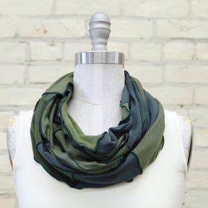 Puede incluir: Bufanda infinita verde y azul exhibida en un maniquí. La bufanda está hecha de tela suave y tiene un diseño en capas. La paleta de colores incluye tonos de verde y azul, creando un accesorio visualmente atractivo. El fondo es una pared de ladrillos blancos.