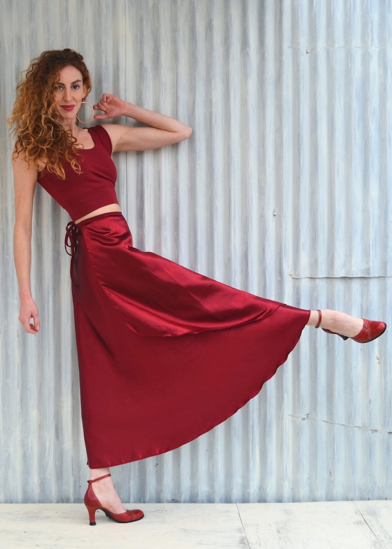 Puede incluir: Una mujer con un top rojo corto y una falda larga de sat&eacute;n rojo que se envuelve. Est&aacute; de pie con una pierna levantada y el pie apuntando, llevando tacones rojos.