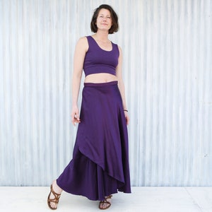 Puede incluir: Una mujer con un top corto morado y una falda larga de envoltura morada. Est&aacute; de pie frente a una pared de metal corrugado blanco.