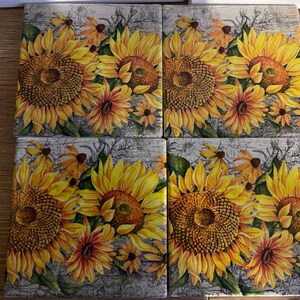 May include: Four square ceramic coasters with a sunflower design. Each coaster features a large yellow sunflower with a brown center and smaller orange flowers. The background has a distressed, weathered look.
