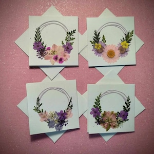May include: Four white cards with a wreath of pressed flowers on each. The wreaths are made of purple, pink, and yellow flowers.