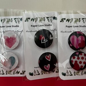 May include: Three sets of heart-themed buttons on white cards, each in a clear plastic sleeve. The buttons feature watercolor hearts, striped hearts, and polka dot hearts. The cards read "Paper Love Studio Handmade Mento Crafts."