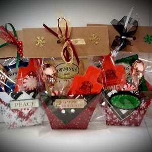 May include: Three Christmas gift bags with red and white patterned fabric baskets filled with tea, candy, and a spoon. The bags are tied with red and green ribbon and have a brown card with a snowflake design.