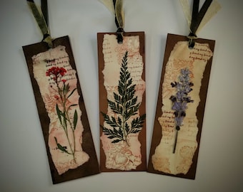 Può includere: Tre segnalibri fatti a mano con fiori pressati e carta decorativa. Ogni segnalibro presenta un fiore pressato diverso: rosso, verde e viola. I segnalibri hanno bordi marroni e sono adornati con nastri.