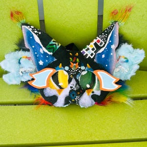 May include: A colorful, handmade cat mask with blue, black, and white accents. The mask features large, expressive eyes, blue ears with white dots, and a rainbow of feathers. The mask is resting on a green surface.