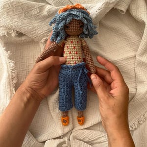 Può includere: Una bambola all'uncinetto fatta a mano con capelli blu e una fascia arancione. La bambola indossa un top beige con pois rossi, pantaloni blu e scarpe arancioni. La bambola è tenuta da due mani.