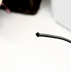 May include: Close-up of a black elastic cord, likely for crafting or sewing projects. The cord is thin and flexible, with a slightly textured surface. The background is white, with a glimpse of other items. The image includes the logo for Jojo's Quilt Shoppe.