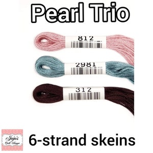 Può includere: Tre gomitoli di filo da ricamo in rosa, blu e marrone. I gomitoli sono etichettati con i numeri 812, 2981 e 312. Il testo "Pearl Trio" e "6-strand skeins" è anche visibile.