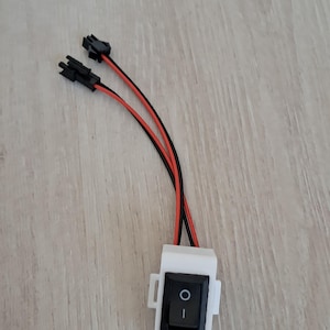 Puede incluir: Un interruptor eléctrico blanco con un botón de encendido/apagado negro y cables rojos y negros. El interruptor es rectangular y tiene dos conectores negros. Los cables están conectados al interruptor.