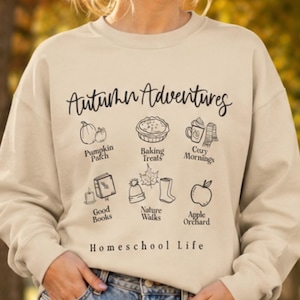 May include: A beige crewneck sweatshirt featuring black line art illustrations and text that reads Autumn Adventures, Pumpkin Patch, Baking Treats, Cozy Mornings, Good Books, Nature Walks, Apple Orchard, and Homeschool Life.