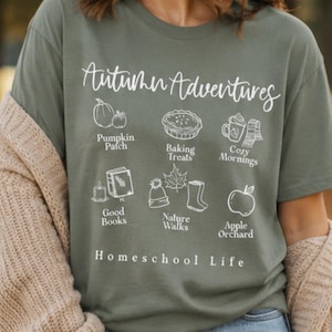 May include: An olive green short-sleeve t-shirt featuring white line art graphics and text that reads Autumn Adventures, Pumpkin Patch, Baking Treats, Cozy Mornings, Good Books, Nature Walks, Apple Orchard, and Homeschool Life.