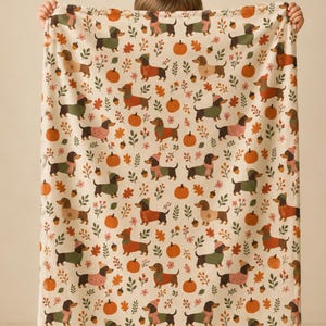 May include: A cream-colored throw blanket featuring a repeating pattern of brown dachshunds wearing colorful sweaters and hats, interspersed with orange pumpkins, brown acorns, and small green leaves, creating a festive autumn-themed design.