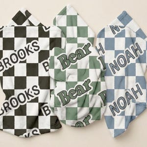 May include: Three soft blankets featuring a checkered pattern. The first is black and white with the name BROOKS, the second is sage green and white with the word Bear, and the third is dusty blue and white with the name NOAH printed in bold letters.