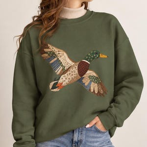May include: An olive green crewneck sweatshirt featuring a large graphic of a flying duck. The duck is designed with a patchwork pattern consisting of various floral and geometric prints in shades of blue, brown, cream, and green with a yellow beak.