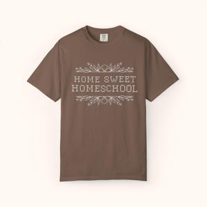 May include: A brown t-shirt with the words "HOME SWEET HOMESCHOOL" in white, framed by floral and leaf designs. The shirt has short sleeves and a crew neck. The text is centered on the front of the shirt.
