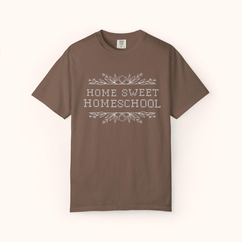 May include: A brown t-shirt with the words "HOME SWEET HOMESCHOOL" in white, framed by floral and leaf designs. The shirt has short sleeves and a crew neck. The text is centered on the front of the shirt.