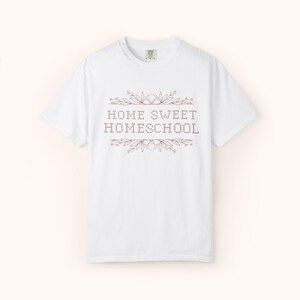 May include: White t-shirt with the text "HOME SWEET HOMESCHOOL" in a brown, pixelated font, framed by floral and leaf designs. The shirt has a crew neck and short sleeves, perfect for casual wear.