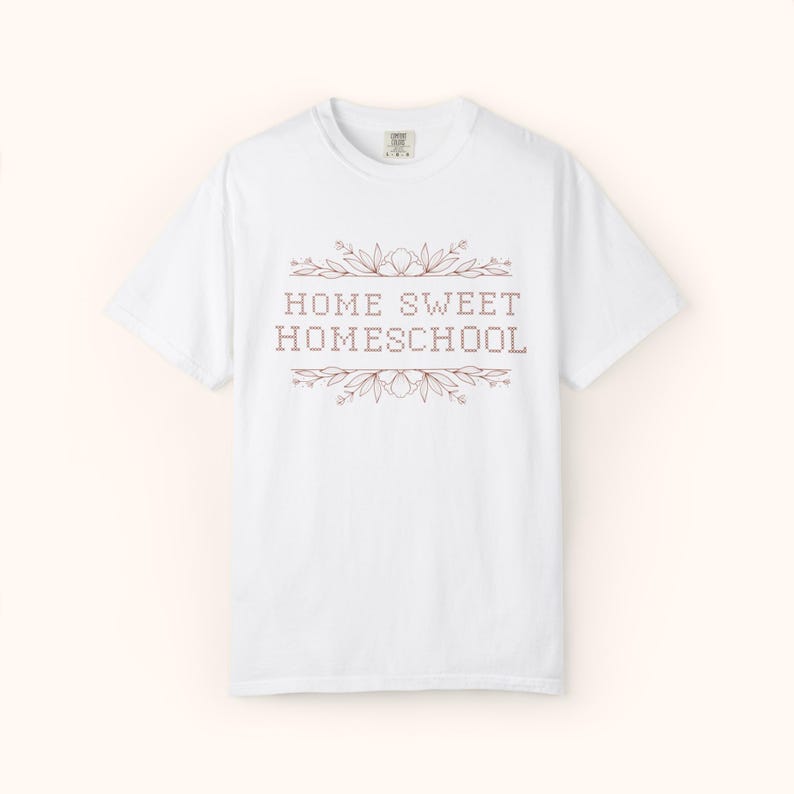May include: White t-shirt with the text "HOME SWEET HOMESCHOOL" in a brown, pixelated font, framed by floral and leaf designs. The shirt has a crew neck and short sleeves, perfect for casual wear.