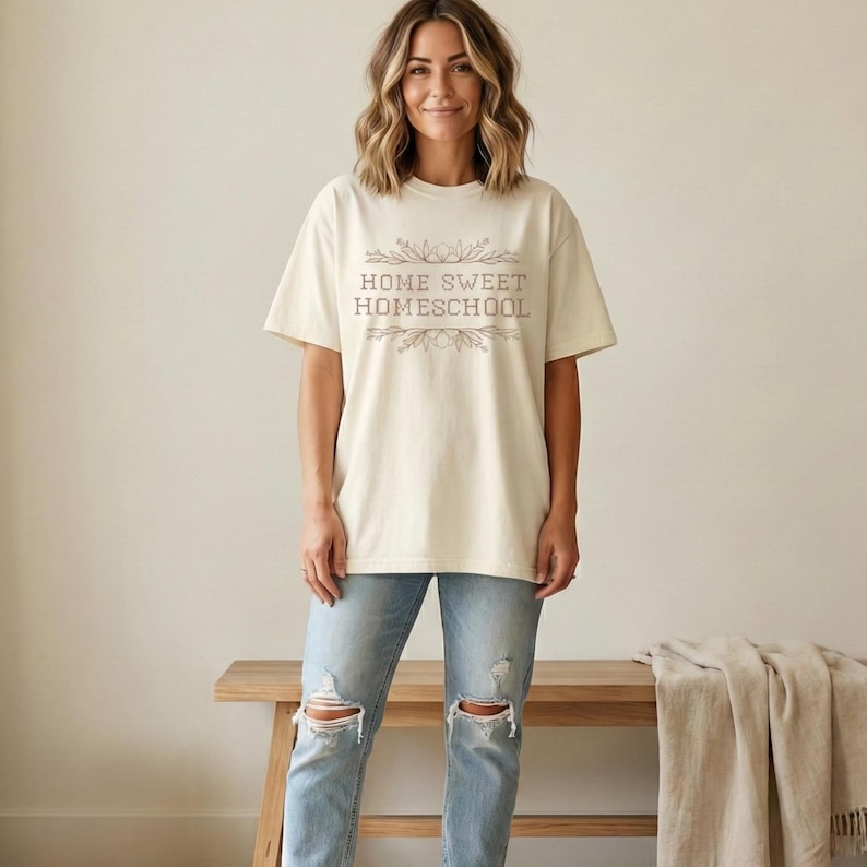 May include: Cream-colored t-shirt with the text "HOME SWEET HOMESCHOOL" in a decorative font. The shirt is paired with light blue jeans. A wooden bench and a beige blanket are in the background.