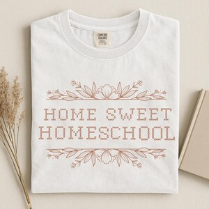 May include: White t-shirt with the text "HOME SWEET HOMESCHOOL" in a brown, cross-stitch style font. The text is framed by floral and leaf designs. The shirt has a crew neck and short sleeves.