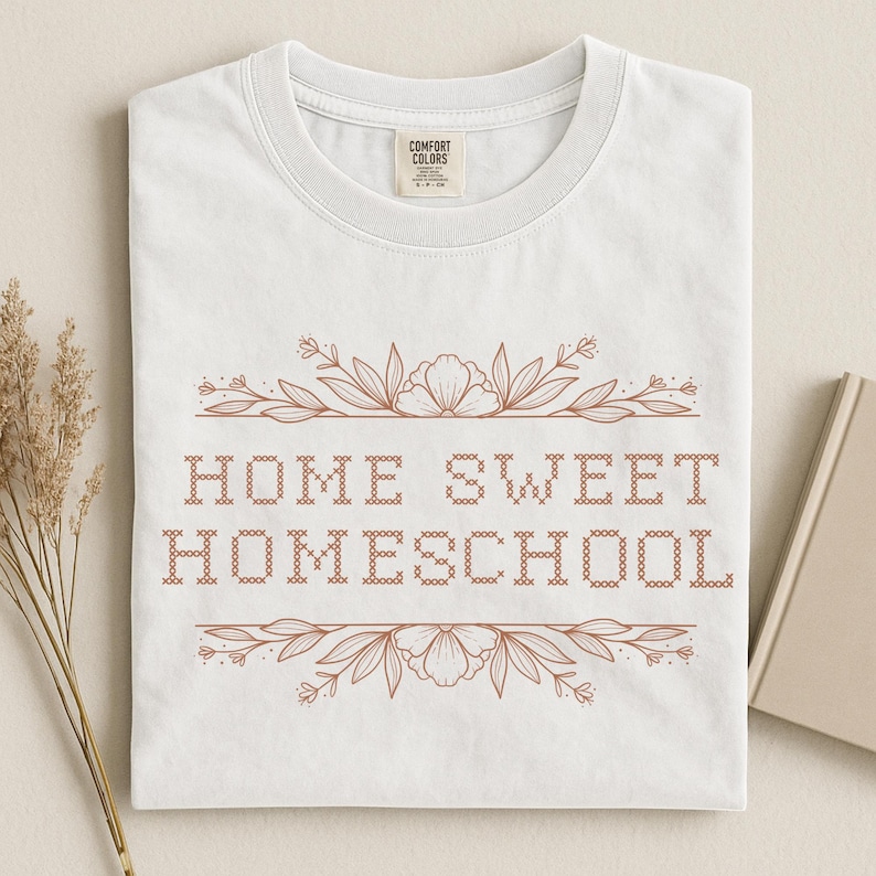 May include: White t-shirt with the text "HOME SWEET HOMESCHOOL" in a brown, cross-stitch style font. The text is framed by floral and leaf designs. The shirt has a crew neck and short sleeves.
