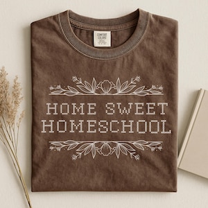 May include: A brown t-shirt with the words "HOME SWEET HOMESCHOOL" in white, surrounded by floral designs. The shirt is folded, with the collar visible. The brand tag "COMFORT COLORS" is visible.