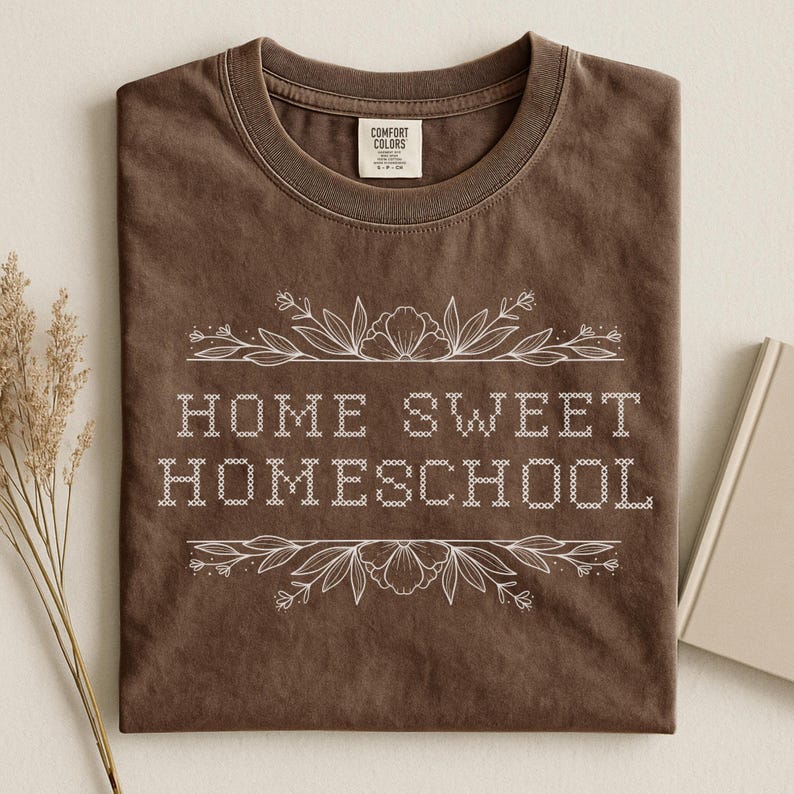 May include: A brown t-shirt with the words "HOME SWEET HOMESCHOOL" in white, surrounded by floral designs. The shirt is folded, with the collar visible. The brand tag "COMFORT COLORS" is visible.