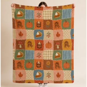 Op de afbeelding: Een rechthoekige plaid met een patchworkontwerp van afwisselende vierkanten in tinten oranje, bruin, blauw en crème. Het patroon bevat illustraties van pompoenen, taartpunten, kaarsen, paddenstoelen, bladeren en sjaals.