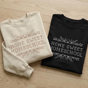 May include: Two sweatshirts, one cream-colored and one black, are displayed. Both feature the text "HOME SWEET HOMESCHOOL" in a decorative font, framed by floral and leaf designs. The cream sweatshirt is a crewneck, while the black one is a t-shirt.