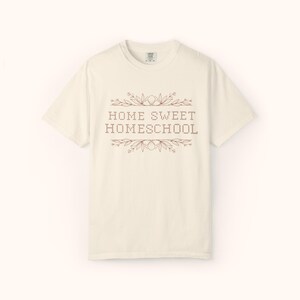 May include: Cream-colored t-shirt with the text "HOME SWEET HOMESCHOOL" in a brown, decorative font. The text is framed by floral and leaf accents. The shirt has a crew neck and short sleeves.