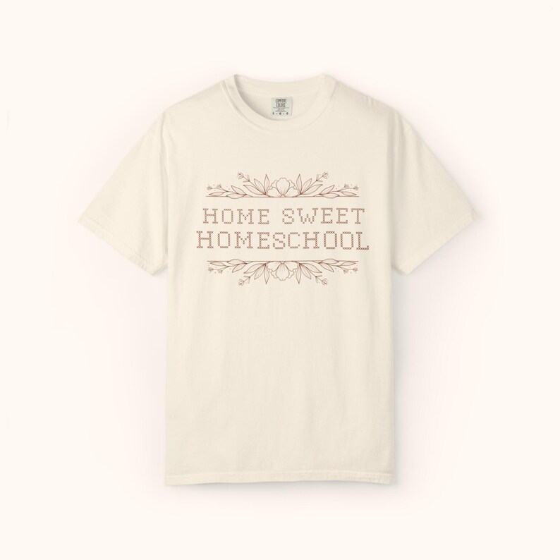 May include: Cream-colored t-shirt with the text "HOME SWEET HOMESCHOOL" in a brown, decorative font. The text is framed by floral and leaf accents. The shirt has a crew neck and short sleeves.