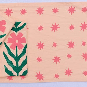 Puede incluir: Un camino de mesa color melocotón y dos servilletas a juego. El camino de mesa y las servilletas están decorados con estrellas rosas y un diseño floral con flores rosas y hojas verdes. Los artículos son de tela.