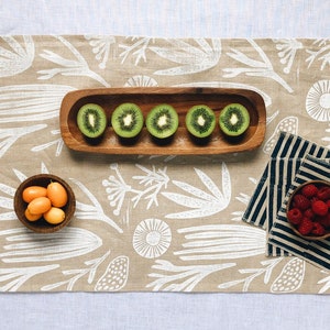 May include: A beige linen table runner with a white floral pattern. The runner is decorated with a wooden bowl of kiwi slices, a wooden bowl of orange kumquats, and a small bowl of red raspberries on a set of black and white striped napkins.