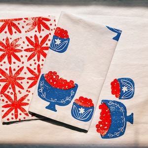 May include: Two white tea towels with red and blue printed designs. One towel features a repeating pattern of red flowers with white dots. The other towel has a design of red berries in blue bowls with white flowers.