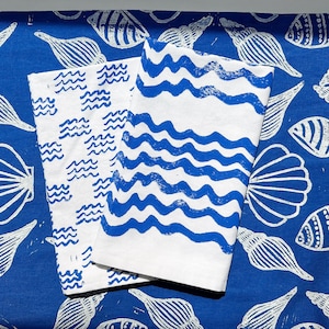 Puede incluir: Dos paneles de tela blanca con patrones azules de ondas y cuadrículas. Los paneles están colocados sobre un fondo de tela azul con un patrón de conchas blancas.