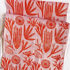 Może przedstawiać: Różowy lniany ręcznik kuchenny z czerwonym wzorem kwiatowym. Wzór przedstawia stylizowane kwiaty, liście i łodygi.