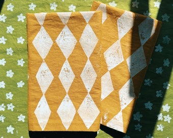 Pode incluir: Duas toalhas de tecido amarelo mostarda com um padrão de diamantes brancos. As toalhas estão dobradas e colocadas sobre um fundo de tecido verde com desenhos de flores brancas.