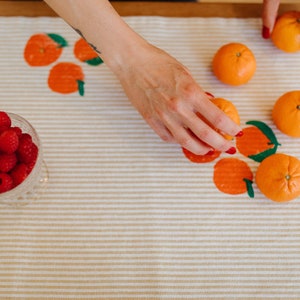 Peut inclure: Une nappe beige à rayures ornée d'illustrations d'oranges. Des mandarines fraîches et un bol en verre de framboises sont disposés sur la table. Une main tend la main vers une mandarine.