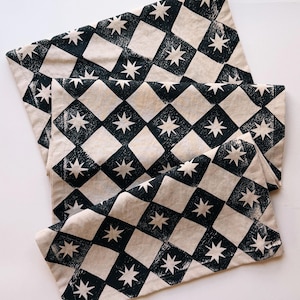 May include: A set of two folded cream-colored fabric napkins with a repeating pattern of black diamond shapes and white starbursts. The napkins are made of a soft, textured material.