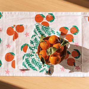 Puede incluir: Un paño de cocina de algodón blanco con un patrón de hojas verdes y fruta cítrica naranja impresa. Un tazón de naranjas frescas se encuentra encima de la toalla.