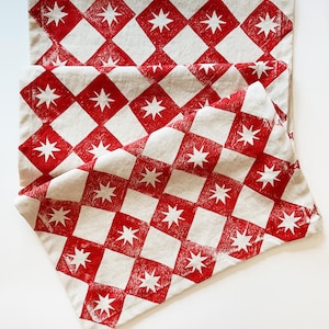 May include: Red and white checkered table runner with a star pattern. The runner is made of linen and has a rustic, farmhouse style.