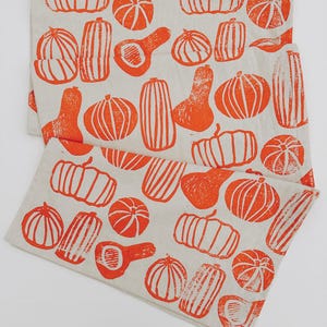 May include: Two folded linen tea towels with a repeating pattern of orange pumpkins in various shapes. The pumpkins are printed on a neutral, off-white background. The design is a block print style, perfect for fall.