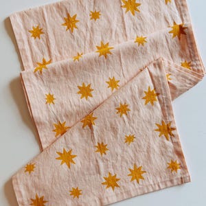 Puede incluir: Un camino de mesa rosa claro con un patrón repetido de estrellas amarillo dorado. Las estrellas, de diferentes tamaños, están distribuidas uniformemente sobre la tela. El camino de mesa está doblado, mostrando el diseño.