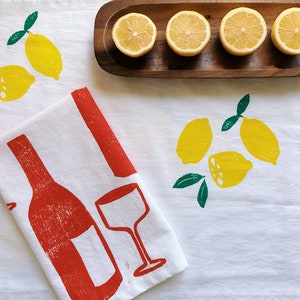 Puede incluir: Una toalla de cocina blanca con un dise&ntilde;o de botella de vino tinto y vaso. La toalla tambi&eacute;n tiene un estampado de lim&oacute;n verde y amarillo. Hay cinco limones en una bandeja de madera.