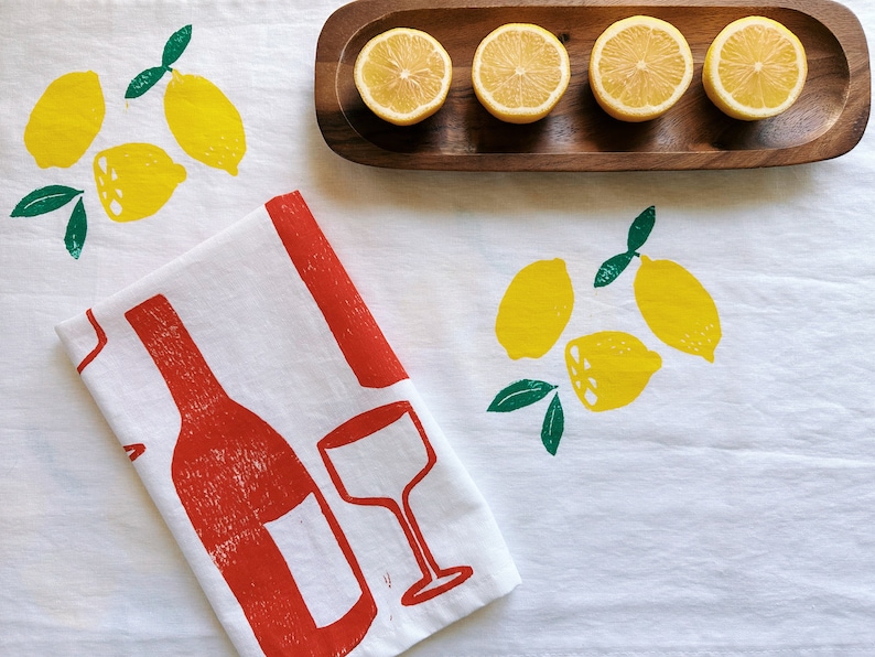 Puede incluir: Una toalla de cocina blanca con un dise&ntilde;o de botella de vino tinto y vaso. La toalla tambi&eacute;n tiene un estampado de lim&oacute;n verde y amarillo. Hay cinco limones en una bandeja de madera.
