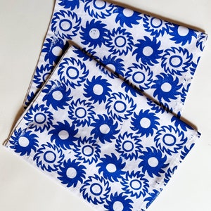 May include: Two white tea towels with a blue and white geometric pattern. The pattern is a repeating design of stylized sunbursts.