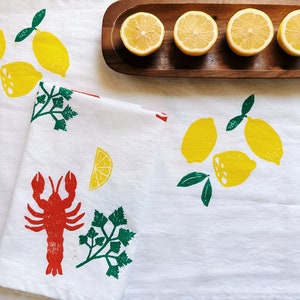 Puede incluir: Un pa&ntilde;o de cocina blanco con un langostino rojo, perejil verde y limones amarillos impresos en &eacute;l. El pa&ntilde;o de cocina est&aacute; doblado por la mitad y est&aacute; sobre un mantel blanco con limones en una bandeja de madera en el fondo.